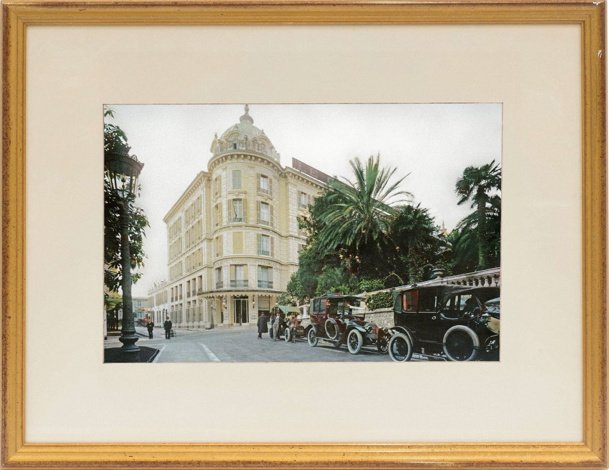 Vintage cars on a street with a large building and palm trees in the background, framed in gold.